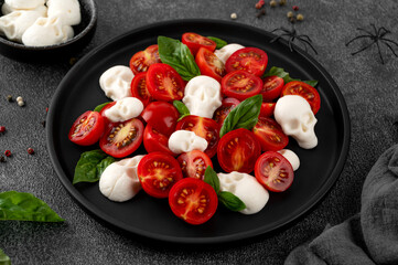 Halloween food. Caprese salad with mozzarella cheese skulls, basil and balsamic dressing on a black plate on a dark background. Funny food idea.