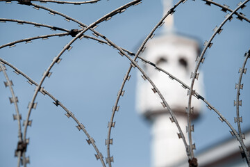 A close-up view of barbed wire, with a blurred tower in the background against a clear blue sky. Suggests themes of security, confinement, or restricted areas in an outdoor setting.