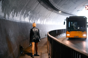 Single woman walks along a pathway in a dimly lit tunnel, as a bus approaches on an adjacent road. The lighting and environment create a modern, urban atmosphere. © Loginova