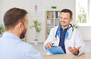 Portrait of a happy smiling young man doctor talking with male patient in office giving...