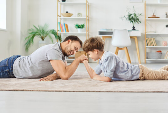 Cheerful father and little son enjoy a friendly arm-wrestling competition game at home. Playful contest of strength highlights family bond and spending leisure time together in a sporty way.