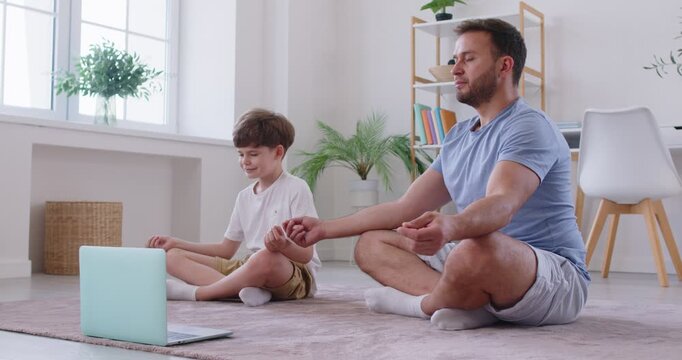 Relaxed family father and son doing yoga breathing exercise, following online video tutorial. Little boy and young man enjoying meditation with closed eyes in lotus pose at home. Family workout.