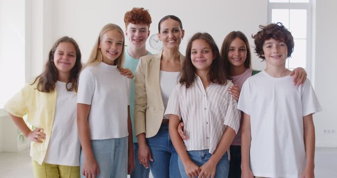 Portrait of cheerful group of happy schoolchildren standing in classroom with their teacher. Friendly Caucasian female teacher stands in middle of group of children in school classroom. 4k video.