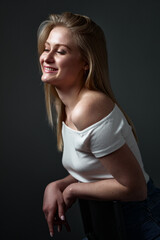 A cheerful young woman wearing a white shirt relaxes indoors against a dark background, smiling...