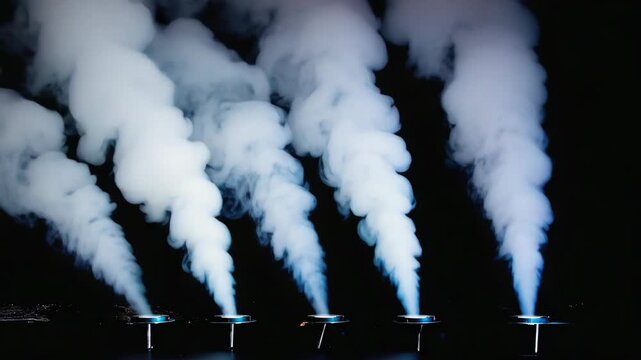 Smoke machines create vertical columns of smoke rising from vents in a stage floor, illuminated with blue light against a black background, suggesting a mysterious or suspenseful atmosphere