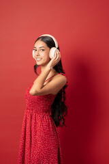 young woman smiling at camera holding headphones in red dress against red background vertical