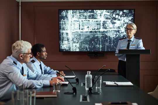 Middle aged Caucasian woman in uniform standing at podium presenting to senior Caucasian man and middle aged Black man in uniforms, discussing aerial map on screen during meeting - Powered by Adobe