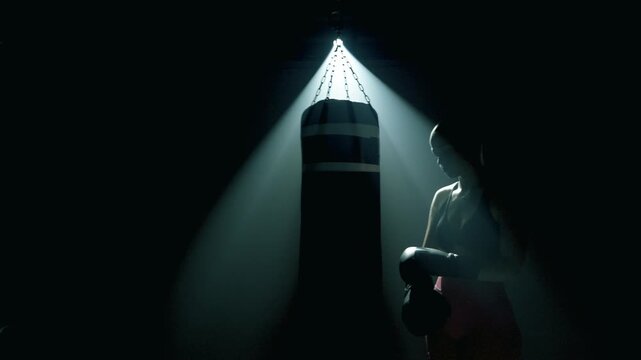 Młoda blondyna zapina sportowe rękawice w ciemnym bokserskim studiu, zbliżenie makro, spięte włosy, dynamiczne oświetlenie, worek treningowy, klimat high‑end fitnessowego wideo