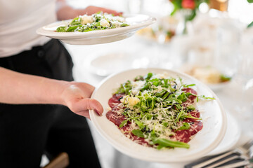 Waiter serving two plates of fresh salad with arugula, grated cheese, and dressing in a bright, elegant restaurant setting. Concept of fine dining and hospitality.