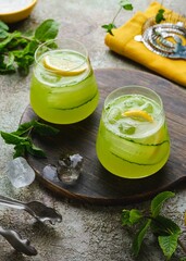 Refreshing drink, cucumber lemonade in glass cups with ice, lemon and cucumber slices on green concrete background. Homemade lemonades.