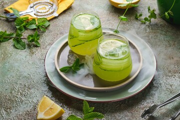 Refreshing drink, cucumber lemonade in glass cups with ice, lemon and cucumber slices on green concrete background. Homemade lemonades.