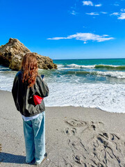 Young brunette woman takes a picture of waves at the shore in Nerja, Spain, gazing at the calm Mediterranean Sea. Peaceful seaside moment filled with connection and serenity.