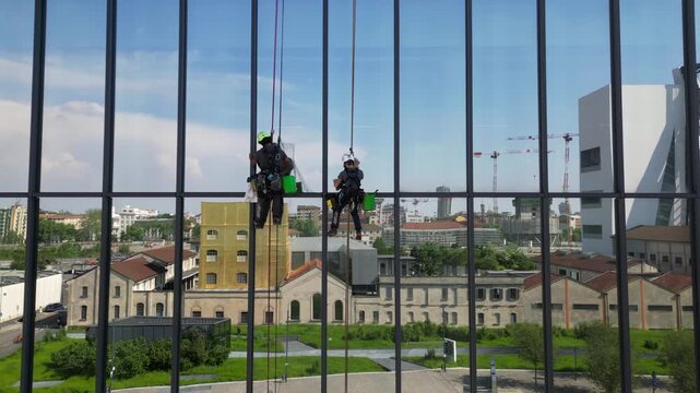Europe, Italy, Milan  07,22,2025 Two skyscraper window cleaners work suspended in the air - drone aerial view of acrobatic window cleaners  near the Prada Foundation new skyline of the fashion city 