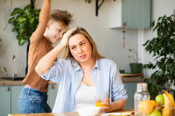 Young woman mom sitting on a kitchen table looking stressed suffers from headache because of noise...