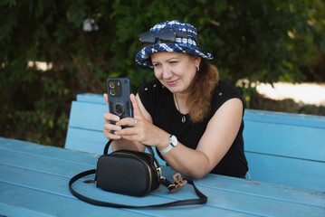 A woman wearing a stylish hat and casual outfit sits at a bench using her smartphone, enjoying the outdoors. A woman relaxes in shade of trees in summer and talks via video call with son