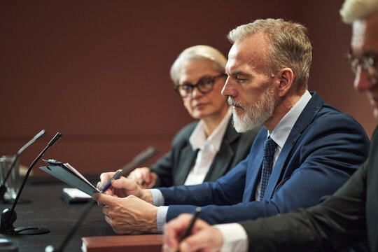 Middle aged Caucasian man reading document at conference table with senior Caucasian woman and middle aged Caucasian man listening, microphones on desk, political meeting setting