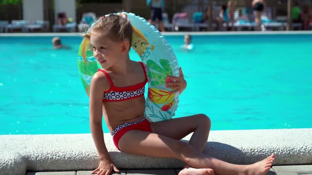 Happy little girl sits by hotel pool with blue water and inflatable ring. Child on summer vacation. Holiday travel