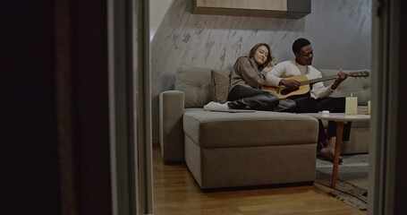 A multiracial couple enjoys a cozy night in, playing guitar and relaxing on the couch by candlelight. The warm atmosphere creates a romantic and intimate setting. - Powered by Adobe