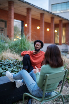 Happy African American male in casual clothes smiling and gesticulating while chilling in yard of apartment building and telling funny story to girlfriend in evening.