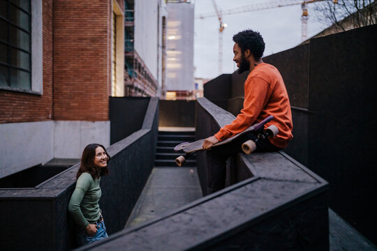 Happy woman with hands in pockets smiling and looking at black male skater while leaning on border outside brick building in evening on city street.