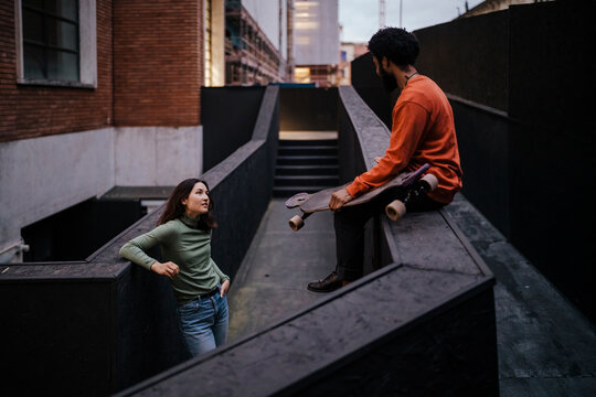 Happy woman with hands in pockets smiling and looking at black male skater while leaning on border outside brick building in evening on city street.