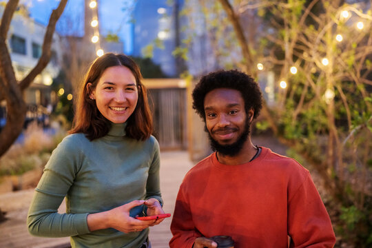 Happy woman with hands in pockets smiling and looking at black male skater while leaning on border outside brick building in evening on city street.