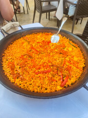 A vibrant serving of traditional Spanish paella, featuring saffron-infused rice, assorted vegetables, and spices, displayed in a rustic pan at a lively outdoor restaurant.
