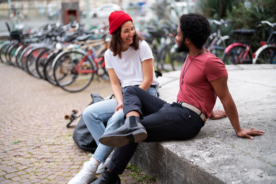 Happy diverse man and woman in casual clothes smiling and looking at each other while sitting on concrete border near bicycles on weekend day in city.