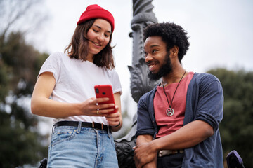From below young woman in casual clothes sharing data in social media with cheerful black boyfriend while speaking weekend day in park.