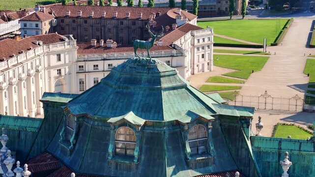 Aerial view of the copper-green roof and deer statue atop the Palazzina di Caccia, set against manicured green lawns and buildings, Stupinigi, Piemonte, Italy.