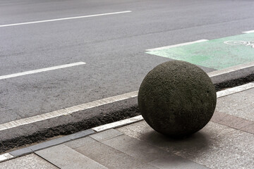 Round stone bollard on a city sidewalk next to an asphalt road with a green painted bicycle lane...