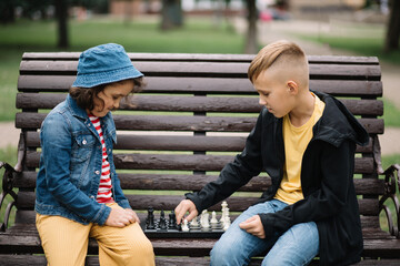 Kids girl and teenager boy playing chess outdoor in a summer park sitting on bench