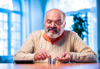 Old Indian man with coin stack and calculator managing money at home table