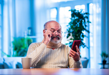 Old Indian man with tea and smartphone browsing content during calm indoor morning