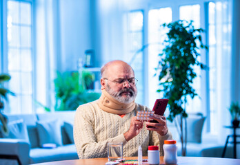 Old Indian man with smartphone and medicine pack verifying ingredients at home