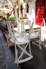 Stacked white chairs on wooden table in a charming street with red door and greenery in the background