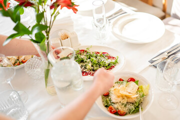 Elegant dining table set with glassware and fresh vegetable salads, one being dressed by hand; vibrant and inviting meal presentation in natural light.