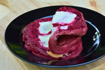 A rich red velvet cake roll, partially unfurled and topped with creamy white frosting, is artfully displayed on a black plate, set against a wooden background