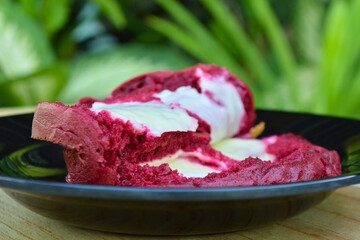 A delicious red velvet cake roll, generously adorned with white cream cheese frosting, is beautifully presented on a black plate with a lush green plant in the soft-focus background