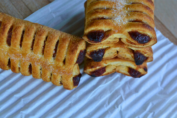 A stack of golden-brown, flaky pastries with visible dark filling, lightly dusted with sugar, stands beside another pastry on a white textured surface, all against a blurred background