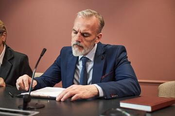 Middle aged Caucasian man sitting at conference table writing on document with pen, wearing suit...