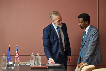 Middle aged Caucasian man and middle aged Black man standing at conference table discussing...