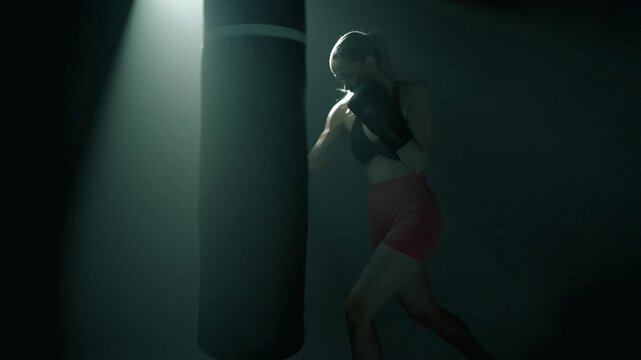 Cała sylwetka bokserki z profilu bijącej w duży worek treningowy w ciemnej sali, jedno filmowe światło podkreśla dramatyzm ruchu, energia, siła, koncentracja, sportowy dramat