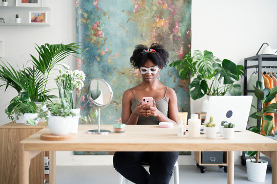 Curly haired black female in sheet facial mask surfing Internet on smartphone and sitting at table at home during beauty routine treatment at home