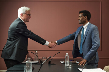Obraz premium Middle aged Caucasian man and middle aged Black man shaking hands across conference table during political meeting, both standing and making eye contact, microphones and water bottles visible