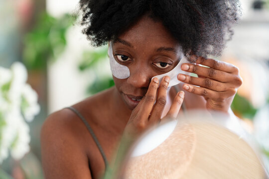 Tranquil black female applying eye patches and looking in round mirror while doing skincare procedure at home