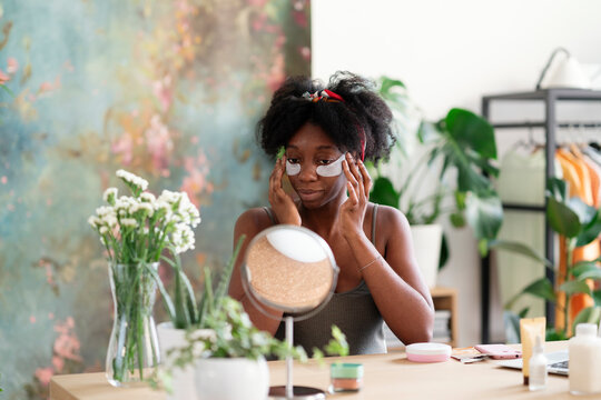 Lovely black female applying hydrating eye patches and looking in round mirror during beauty procedure at home