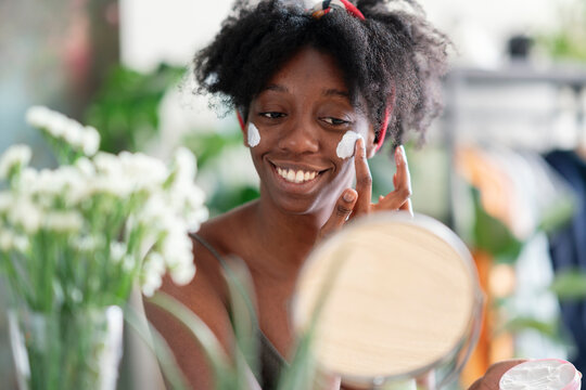 Joyful black female with charming smile applying hydrating face cream and doing beauty routine at home