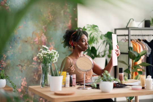 Charming black female sitting at table and doing makeup while looking in mirror