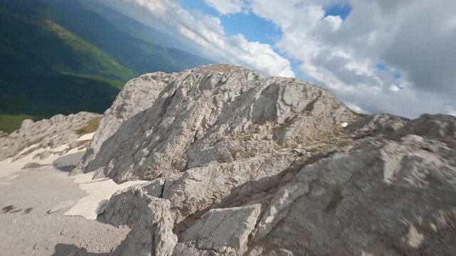 Aerial view of the rugged Komovi Mountain Range with snow patches contrasting vibrant green slopes under a dramatic sky, Комови, Andrijevica Municipality, Montenegro.